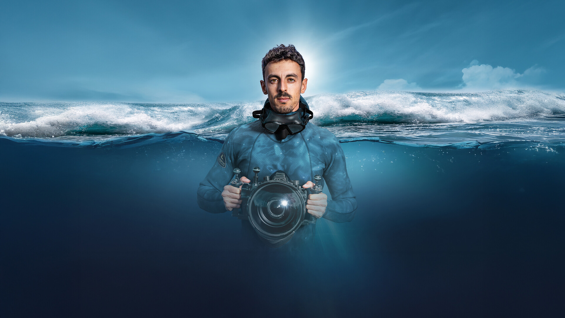 Diver with a diving mask and underwater camera in the ocean, half-submerged, with a breaking wave in the background.