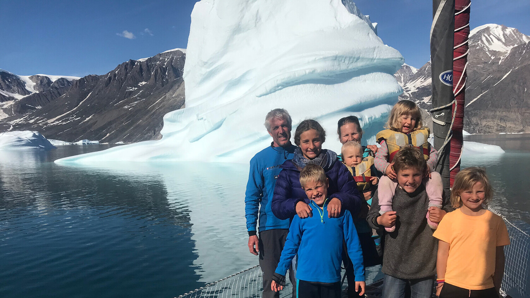 The Schw&ouml;rer family posing together on the deck of their TOPtoTOP expedition sailboat in front of a towering iceberg in a Greenland fjord &ndash; parents and six children on a climate awareness voyage